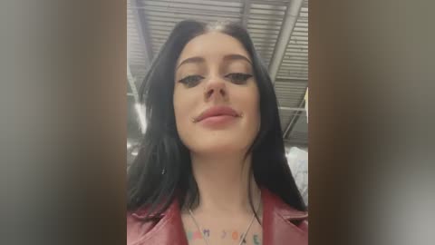 Media: Video of a young woman with long black hair, wearing a red leather jacket, black eyeliner, and a necklace, standing indoors with a metallic ceiling.