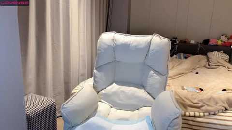 Video of a light beige, plush recliner chair in a cluttered, unmade bedroom with beige walls, a bed with beige bedding, and a small black-and-white checkered ottoman.