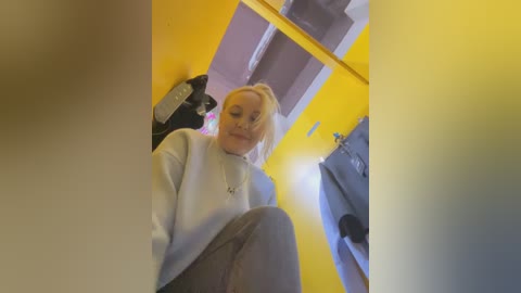 Media: Video of a young woman with blonde hair and pale skin, wearing a light blue sweater and grey pants, sitting in a room with yellow walls, a mirror, and a black jacket hanging on a hook.