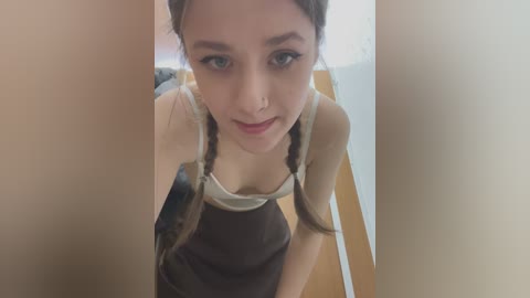 Media: Video of a young Asian woman with light skin, brown hair in pigtails, and green eyes, wearing a tank top and a dark skirt, standing between wooden pillars, indoors, looking directly at the camera.