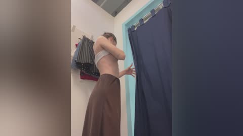 Media: Video of a woman in a dressing room, wearing a white bra and brown skirt, adjusting a navy blue curtain.