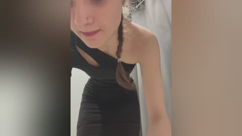 Video of a fair-skinned woman with long brown hair in a braid, wearing a black, strapless dress, leaning forward, and looking up. The background features a beige wall and a blurred, dark object.