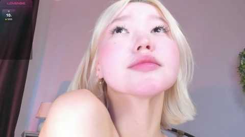 Media: Video of a young, fair-skinned woman with platinum blonde hair, wearing makeup, looking upwards with a pout. Background includes a white wall and a dark curtain.