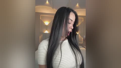 A video of a young woman with long, straight black hair, wearing a cream and white striped top, standing indoors with a modern, minimalist background featuring hanging pendant lights.