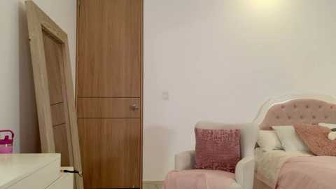 Media: A video of a minimalist, pastel-colored bedroom with a light wooden door, a plush pink armchair, and a white bed with pink and white bedding.