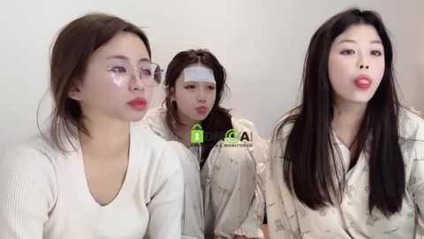 Media: Video of three young Asian women with fair skin, wearing white sweaters and makeup, including face masks, in a minimalist room.