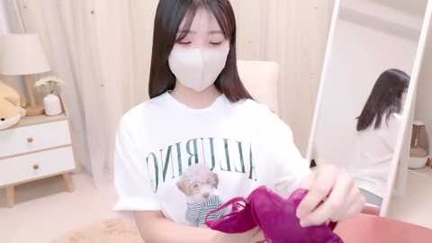 Media: Video of an East Asian woman with long dark hair wearing a white mask, white shirt, and purple gloves, holding a kitten in a cozy, bright bedroom.