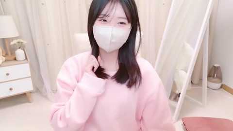 Media: Video of an Asian woman with long black hair, wearing a white face mask, pink sweater, and white socks, sitting in a minimalist bedroom with white walls, a mirror, and a nightstand with a lamp.