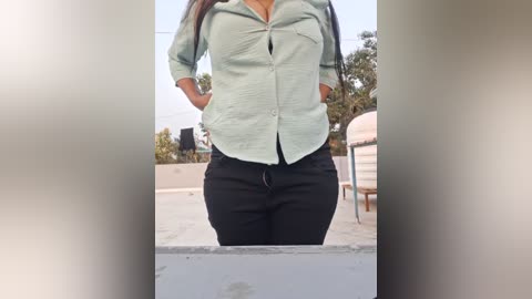 Media: Video of a woman in a light green button-up shirt and black pants, standing outdoors with a blurred background of trees and a metal structure.