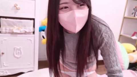 Media: Video of an East Asian woman with long black hair, wearing a gray sweater, white mask, and pink underwear, kneeling on a bed, with a white dresser and stuffed animals in the background.