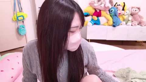 Media: A video of a young Asian woman with long black hair, wearing a grey sweater, sitting on a bed. The room is filled with plush toys, including Minnie Mouse, Mickey Mouse, and a blue bear.