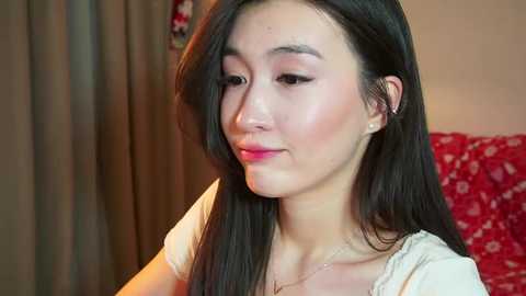 Video of an Asian woman with long black hair, light skin, wearing a white blouse, and subtle makeup, sitting in a room with red-patterned fabric in the background.