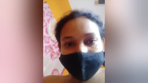 Media: Video of a South Asian woman with medium skin tone, wearing a black mask, black hair, and a pink floral-patterned blouse.