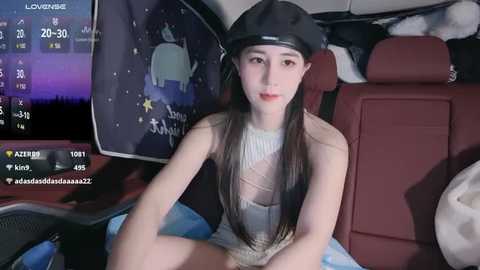 Media: Video of a slim, fair-skinned woman with long black hair, wearing a black beret and a sheer top, seated in a car, looking at the camera.