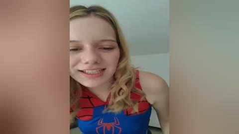 Video of a smiling young woman with long blonde hair, wearing a Spider-Man costume, standing in a minimalist room with beige walls.