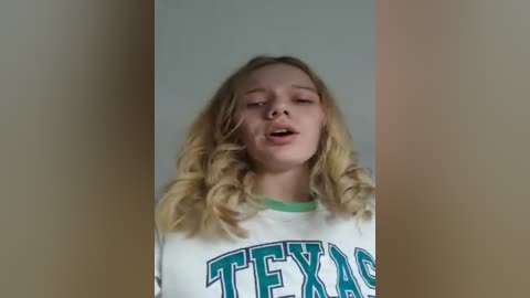 Video of a young Caucasian girl with long, wavy blonde hair, wearing a white Texas University t-shirt, singing with eyes closed against a plain, light beige wall.