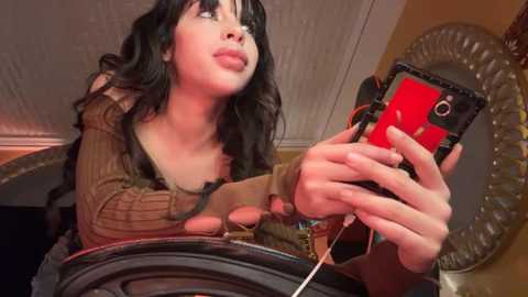 Media: Video of a young woman with long, wavy black hair, wearing a brown sweater, looking at a red phone with a black case, holding a white earbud, in a dimly lit room with a mirror and a decorative wall.