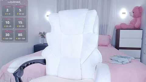 Media: A video of a pastel-pink, plush, high-back gaming chair with a white frame, positioned in a minimalist, girly bedroom with a white bed, a pink teddy bear, and a calendar on the wall.