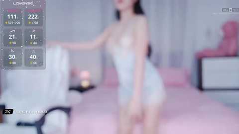 Media: A blurry video of a slender, East Asian woman with long black hair, wearing a revealing white lace lingerie, standing in a pink bedroom with a bed, dresser, and pink chair.