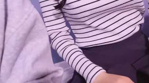 Video of a woman in a white and black striped sweater, dark blue pants, and a gray blanket, with her hand extended towards the camera, against a soft purple background.