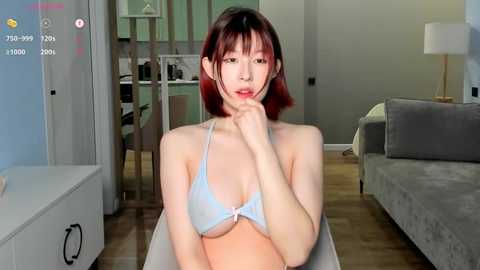 Media: A video of an East Asian woman with short, straight red hair, fair skin, and a petite physique, wearing a light blue lace bra, sitting in a modern living room with wooden floors and a grey sofa.