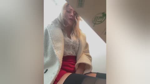 Media: Video of a blonde woman in a white coat, red skirt, and black stockings, sitting in a dimly lit stairwell with a green sticker on the wall.