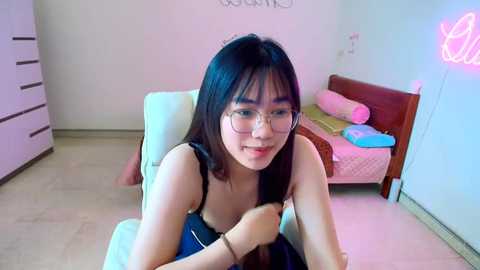 Media: Video of a young Asian woman with straight black hair, glasses, and a blue top, sitting on a bed in a pastel-pink room with a neon sign and a wooden dresser.