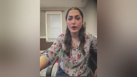 Media: Video of a young woman with long dark hair, wearing a floral blouse, sitting indoors with a window and blinds in the background.