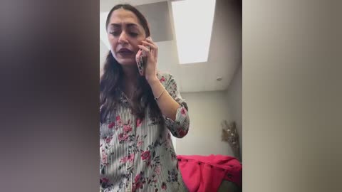 Media: Video of a young woman with long brown hair, wearing a floral blouse, talking on a phone in a modern, brightly lit room with white walls and a ceiling light.