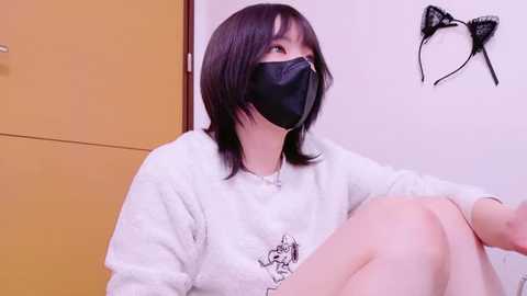 Media: Video of an Asian woman with shoulder-length dark hair, wearing a black face mask and white sweater, sitting against a yellow door, with a black cat head wall decoration.