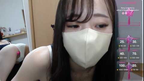 Media: Video of a young East Asian woman with long black hair, wearing a white face mask, sitting on a bed, with a virtual reality overlay showing health stats.