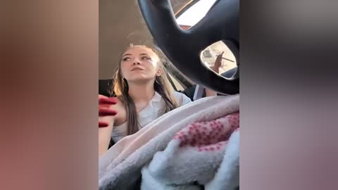 Media: Video of a young woman with long brown hair, wearing a white shirt, sitting in a car. She looks out the window, her red-painted fingernails visible.