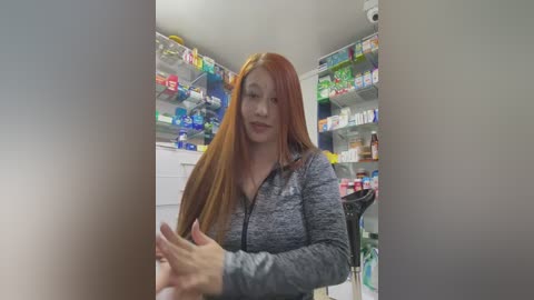 Media: Video of a red-haired woman with fair skin, wearing a gray zip-up jacket, standing in a brightly lit pharmacy with shelves full of medicine bottles.