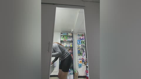 Media: A video captures a woman in a gray long-sleeve top and black shorts bending over to open a refrigerator door. The interior reveals a well-stocked fridge with various colorful items.