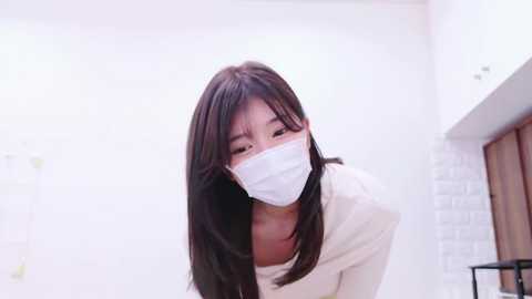 Media: Video of an East Asian woman with long black hair, wearing a white face mask and beige sweater, leaning forward in a modern, white-tiled bathroom with a wooden countertop.