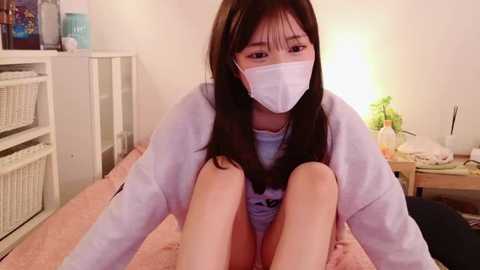 Media: Video of an East Asian woman with long black hair and a white face mask, wearing a light-colored sweater, sitting on a bed with pink sheets, in a cozy bedroom with a white bookshelf and a nightstand.