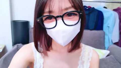 Media: Video of an Asian woman with shoulder-length, straight, dark brown hair, wearing black-framed glasses and a white face mask, a white lace tank top, and sitting on a gray couch. Background includes hanging clothes and a black cylindrical object.