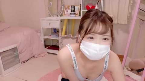 Media: Video of an East Asian woman with light skin, wearing a white face mask, light blue tank top, and black leggings, kneeling on a pink rug in a pastel-colored room with a vanity and bed.