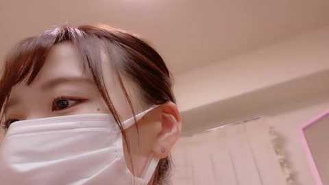 Media: Video of an Asian woman with long brown hair, wearing a white surgical mask, and a small earring in her left ear, indoors with a blurred, light-colored background.