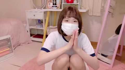 Media: Video of an Asian woman with short brown hair, wearing a white T-shirt, face mask, and seated on a pink rug in a cluttered, feminine bedroom with a vanity mirror and shelves.