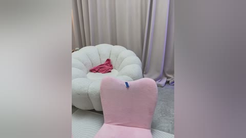 Video of a plush, pink chair with a blue label, situated in a room with cream-colored curtains and a pink blanket draped over a white, tufted pouf.