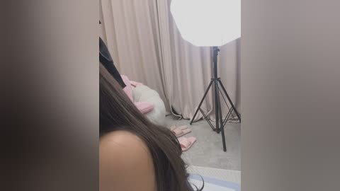 Media: Video of a woman with long brown hair, wearing a black top, sitting on a beige carpet in a room with beige curtains and a black tripod-mounted light.