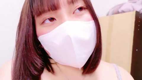 Media: Video of a young Asian woman with straight brown hair, wearing a white face mask, light purple spaghetti strap top, and looking upwards, partially out of focus background.