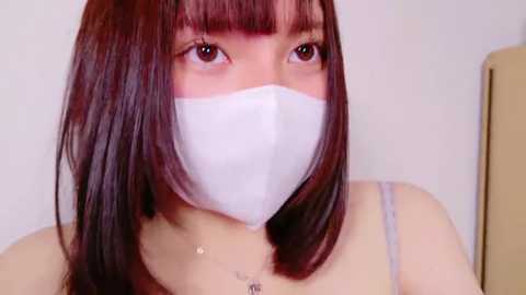 Video of an Asian woman with straight, shoulder-length dark brown hair, wearing a white surgical mask, light gray tank top, and a necklace, against a plain white background.