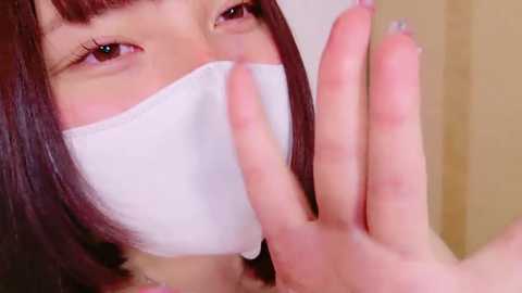 Media: A close-up video of an Asian woman with straight, dark brown hair and fair skin wearing a white surgical mask. She is raising her right hand, displaying neatly manicured nails with a pale pink polish. The background is a blurred beige wall.