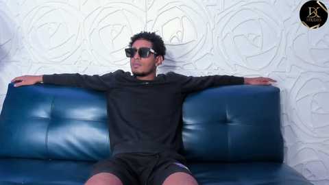 Media: Video of a young Black man with short curly hair, wearing black sunglasses and a long-sleeve shirt, lounging on a blue leather couch against a white, floral-patterned wallpaper.