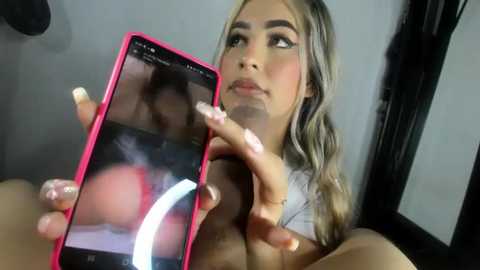 Media: Video of a young woman with long blonde hair and fair skin, wearing a white top, holding a pink smartphone showing a mirror selfie of her breasts.