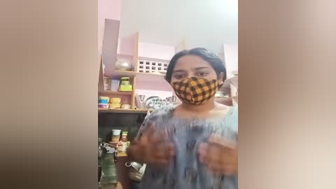 Media: A video of a woman with medium skin tone, wearing a blue shirt and a yellow checkered mask, standing in a cluttered kitchen.