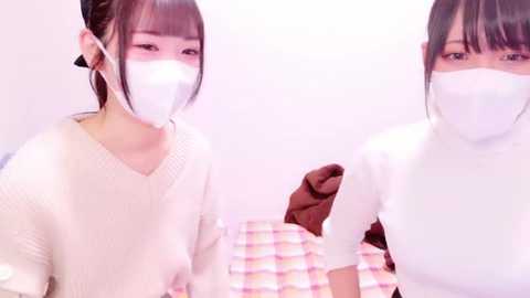 Media: Video of two young Asian women with pale skin and dark hair, wearing white face masks, white sweaters, and sitting on a pastel plaid bedspread. The background is minimalistic with a white wall.