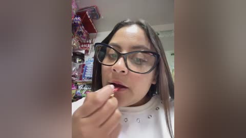 Video of a young Latina woman with long dark hair, glasses, and a white shirt with polka dots, licking a lollipop in a cluttered room with snacks and candy bags.